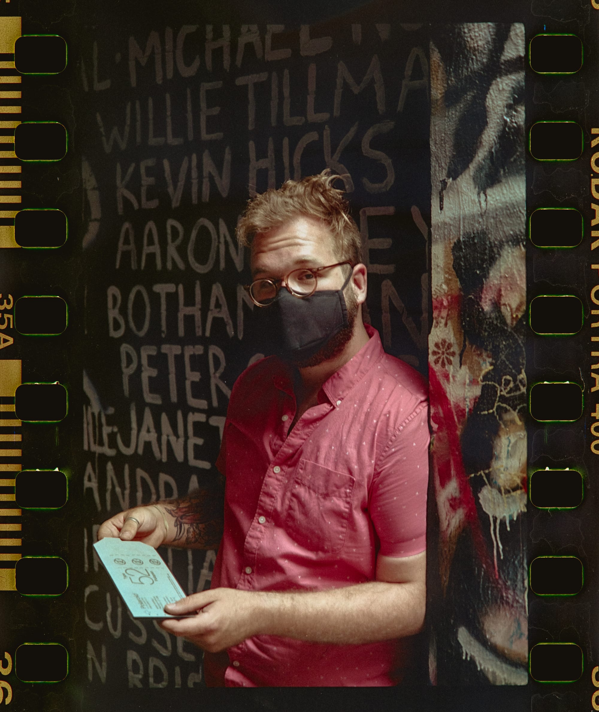 A young white man in a mask in front of a Black Lives Matter mural listing the names of Black people killed by police violence. He's wearing a pink shirt and looking at the camera, holding Polaroid Type 52 pack film in his hands.